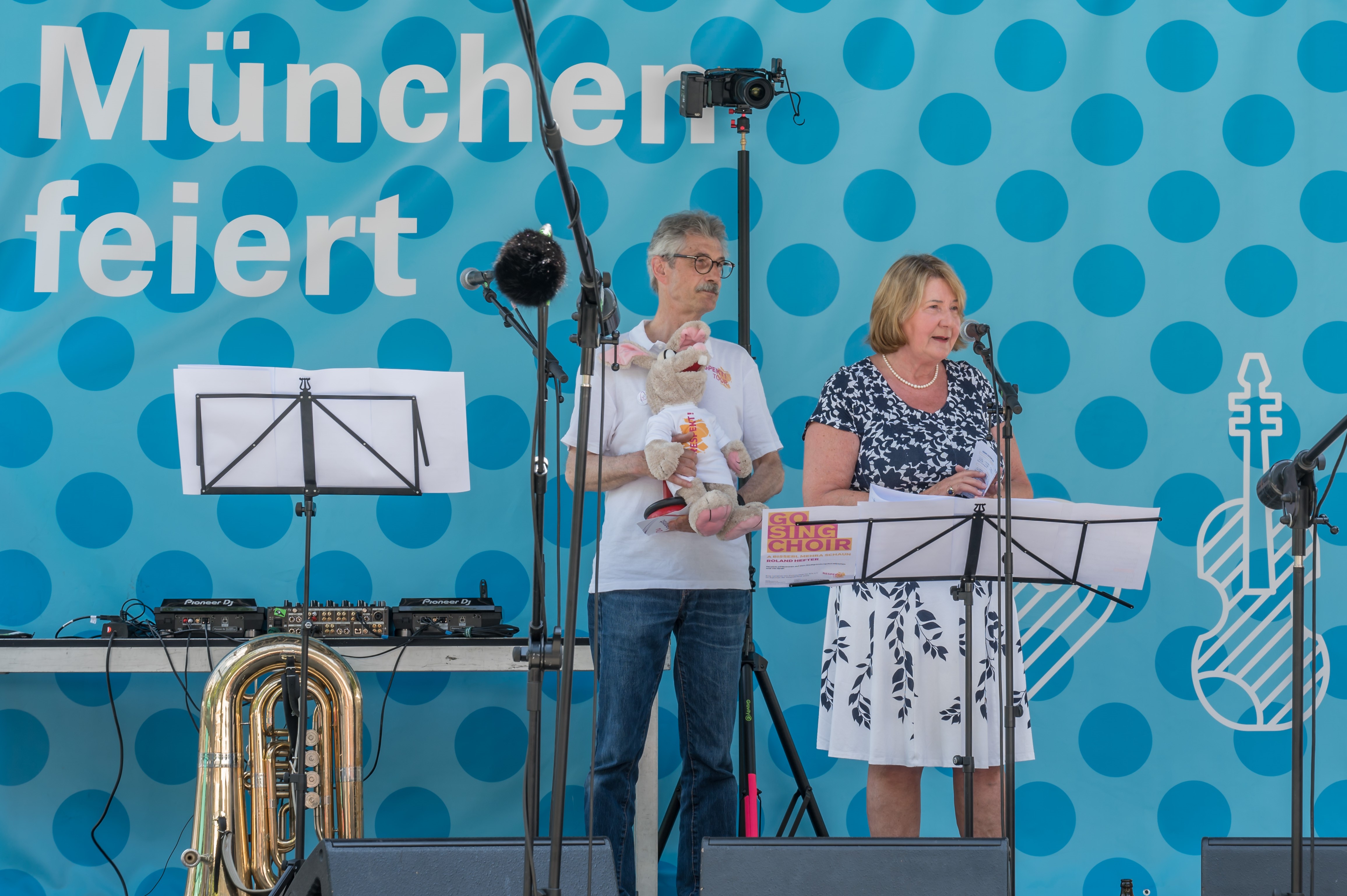 Zwei Personen, eine Bühne, im Hintegrund ein blaues Banner mit der Aufschrift München feiert