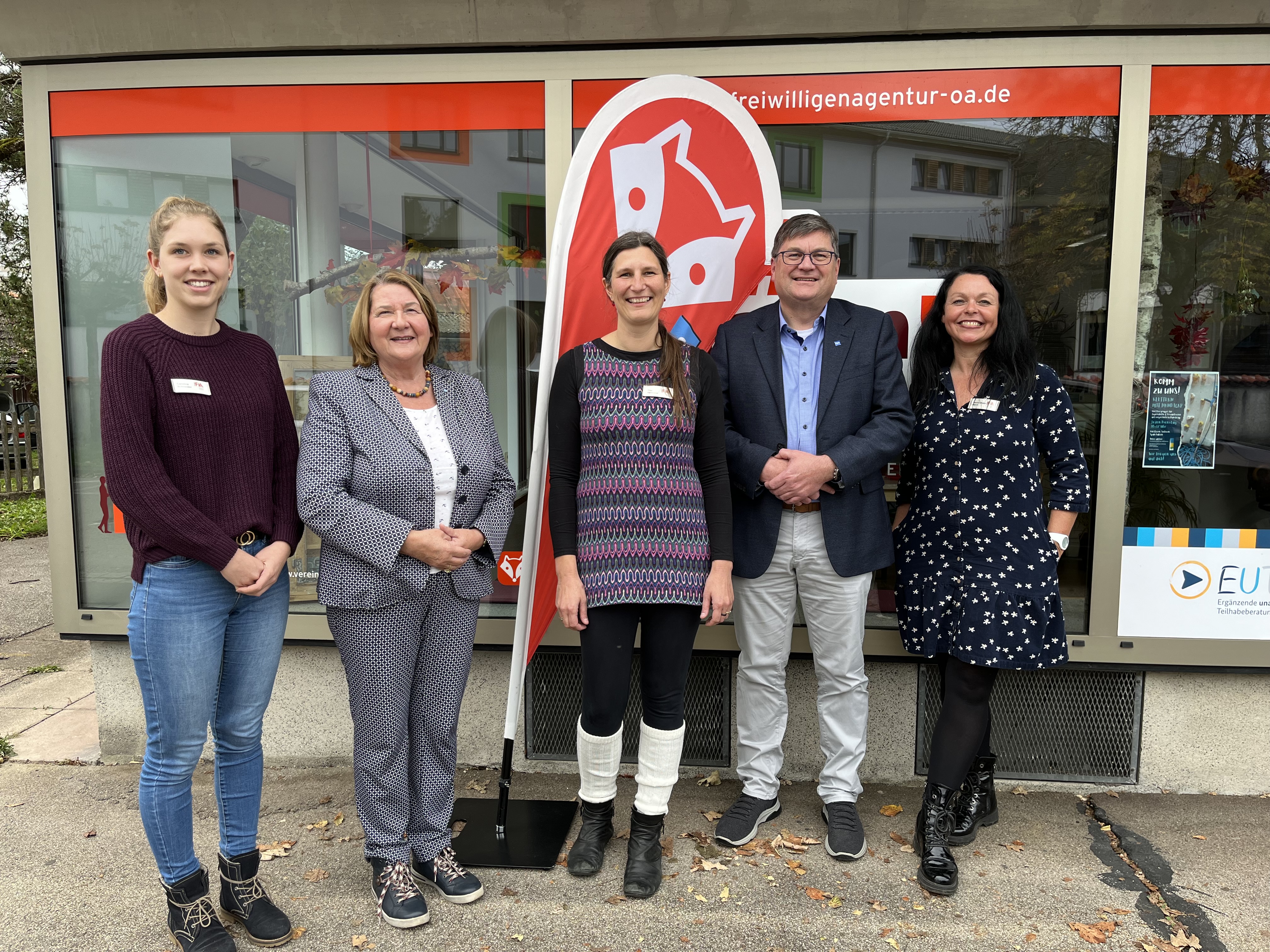 Fünf Personen schauen in die Kamera, im Hintergrund Schaufenster und eine rot-weiße Standfahne