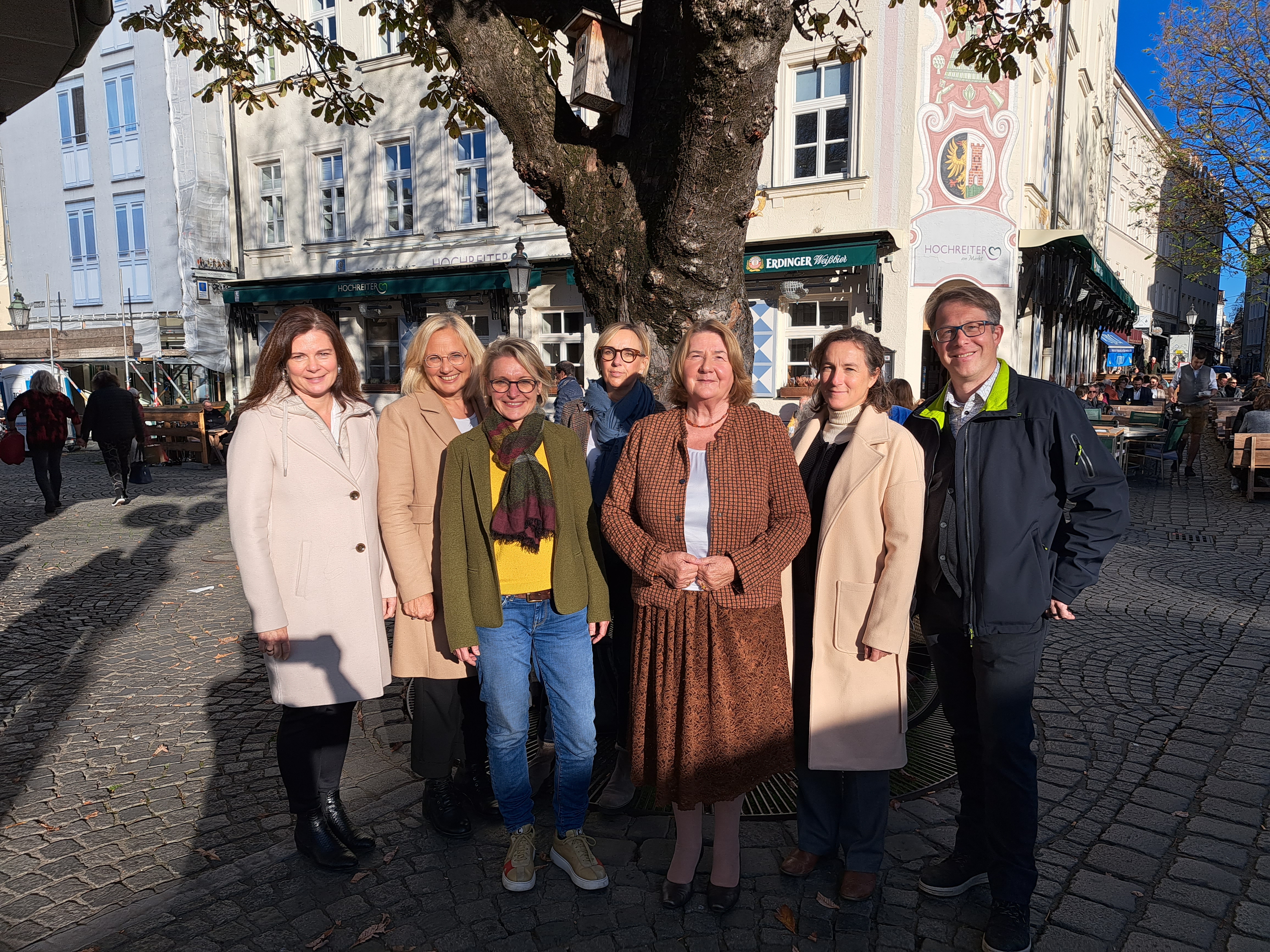 Sieben Personen nehmen vor einem Baum am Münchner Viktualienmarkt Aufstellung, Sonne schein ihnen ins Gesicht, herbstliche Atmosphäre