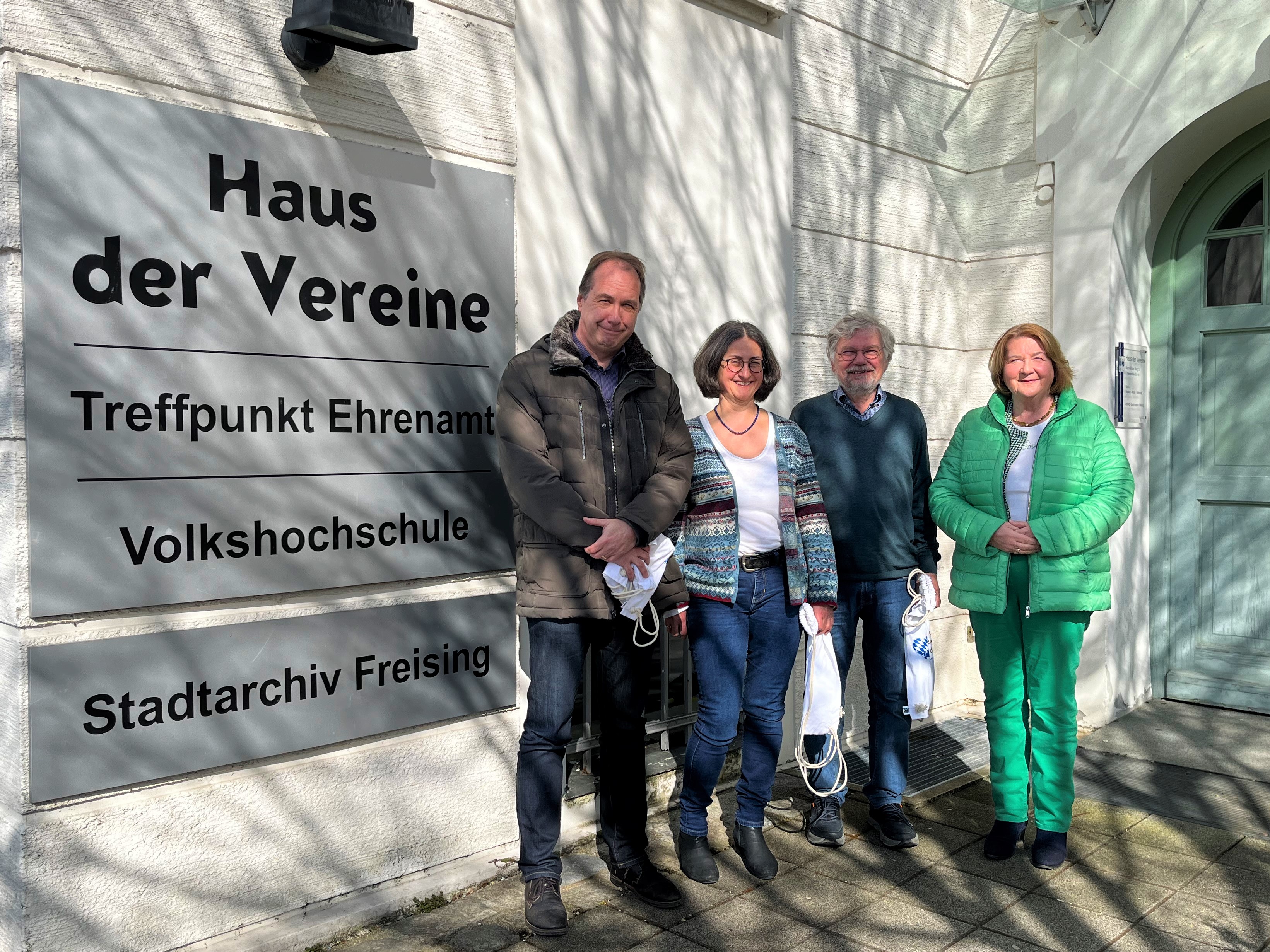 Vier Personen vor einer Hauswand links daneben angebrachte Tafeln, unter anderem mit der Aufschrift Haus der Vereine